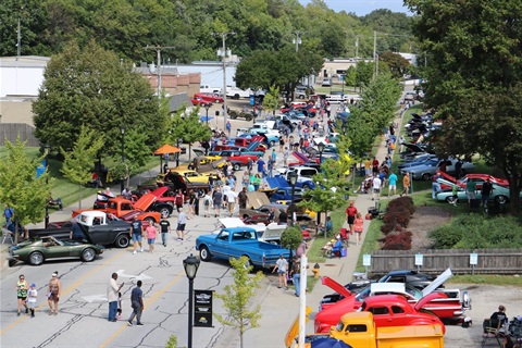 A wide, overhead shot of dozens of colorful cars and trucks from the 1940s and newer. People are walking in-between two rows of cars.