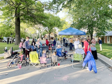 A large group of kids and adults enjoying a block party. A gray stroller sits to the left of the group and there's a tent with a blue cover in the middle of the image.
