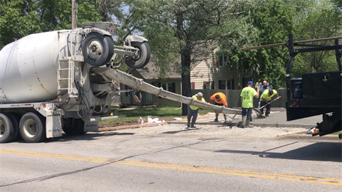 Wideshot image of construction crews pouring concrete onto a driveway. A concrete truck is on the left with four guys on the right.