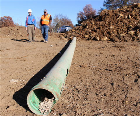 Two men walking at a construction site with a large pipe in foreground. 