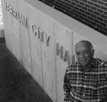 A black and white photo of Mayor Carl Wilkes standing in front of a Merriam City Hall sign on a concrete background. Wilkes, a Black man, is wearing a plaid shirt.