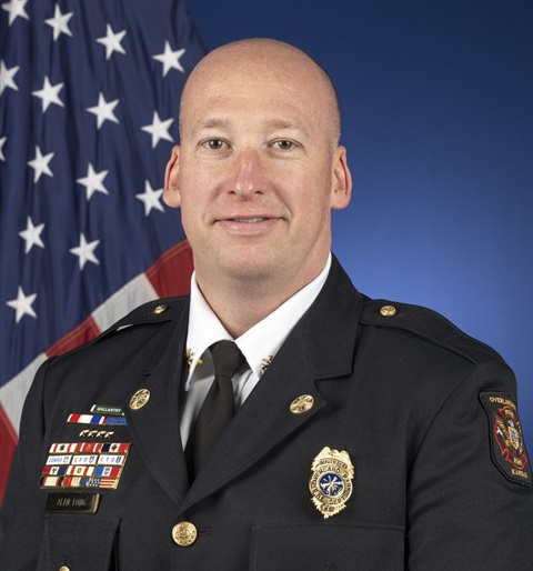 Alan Long, a bald, middle-aged white man, is dressed professionally with a badge and medals on his jacket. He's smiling at the camera for a head shot and an American flag is in the background.