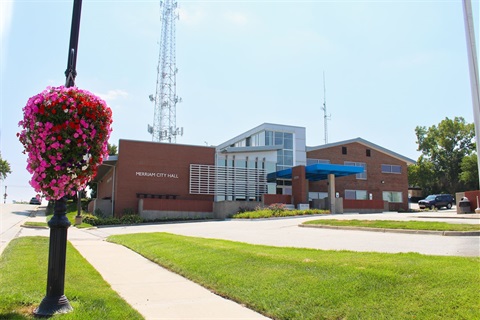 Merriam City Hall is a brick building with a blue overhang. There is a flower basket in the foreground to the left.