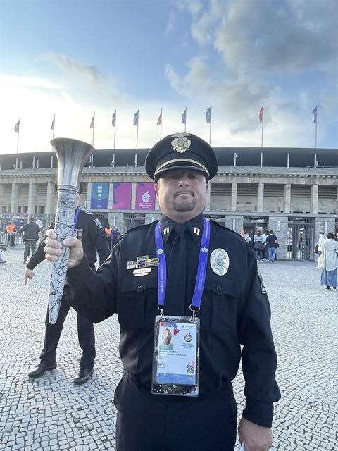 Jeremiah Waters is in full uniform and holding a Torch in his right hand. He's wearing a police hat and standing in front of the entrance to the Special Olympics World Games stadium.