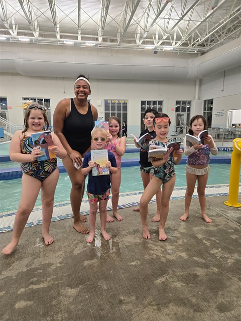 An instructor and kids are holding up book in front of the indoor pool.