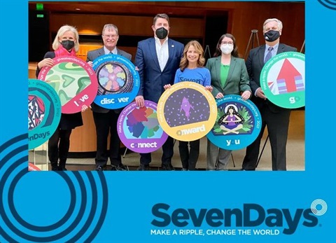 an image of mayor Bob Pape holding a circular board in an emerald green color with a red arrow pointing up. He is standing to the left with five other city leaders and a blue Seven Days logo in front. 