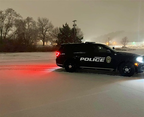A Merriam police vehicle is on a snowy street with its break lights on that is illuminating on the snow.